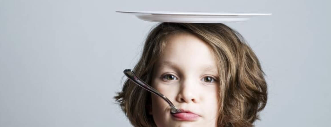 Young girl with a spoon in her mouth balancing a plate on her head