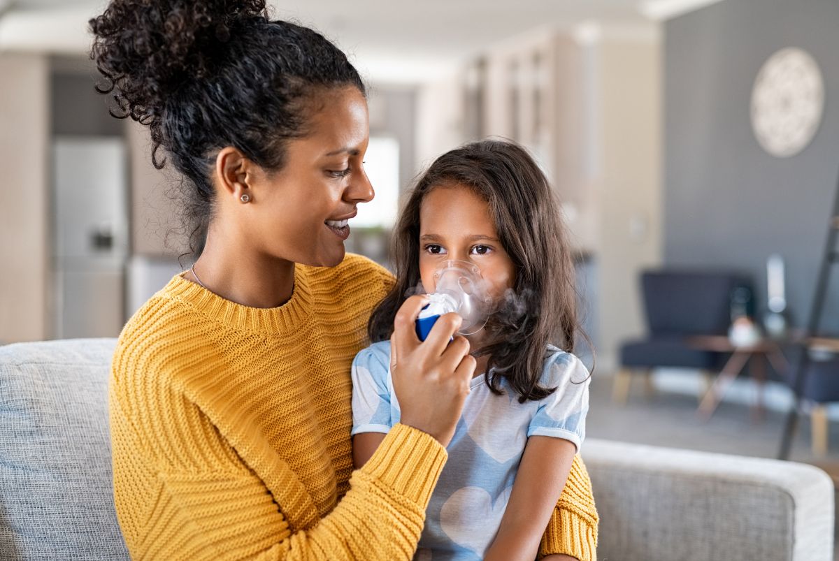 Mother with yellow knit jumper helping daughter with inhaling through a nebulizer. 