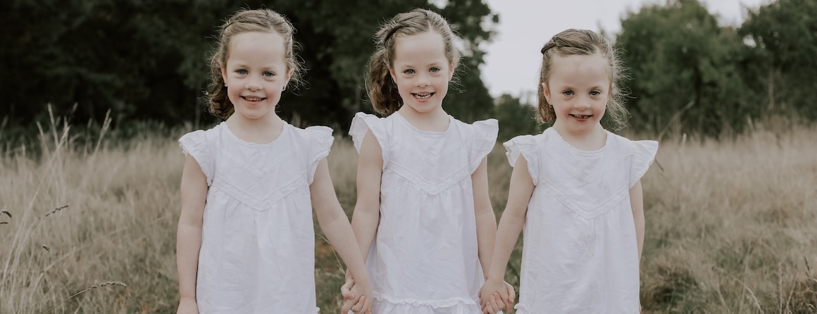 Three young girls in white dresses holding hands in a field. Heart patient and BIGW token designer Emily and her sisters.