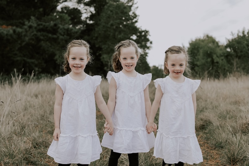 Three young girls in white dresses holding hands in a field. Heart patient and BIGW token designer Emily and her sisters.