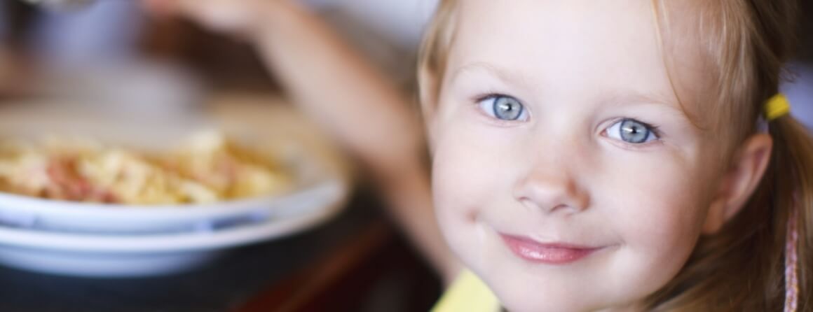 Smiling child looking towards camera with a plate of food in the background.